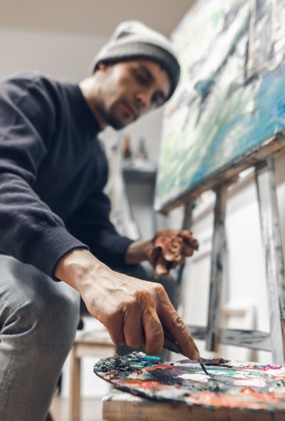 Portrait of a creative man in an art studio at home, creates a picture of oil. Professional artist is painting a picture, mixing oil paint on a palette. Focus on hand with paint and a knife palette