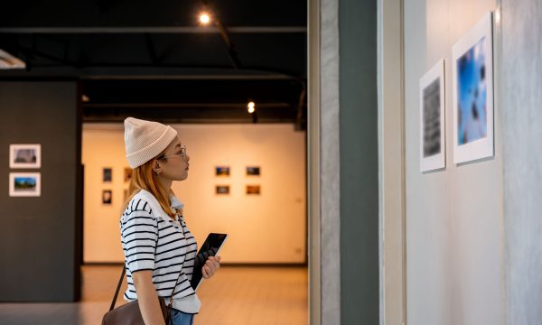 Young person at photo frame hold digital book leaning against at show exhibit artwork gallery, Asian woman holding tablet at art gallery collection in front framed paintings looking pictures on wall