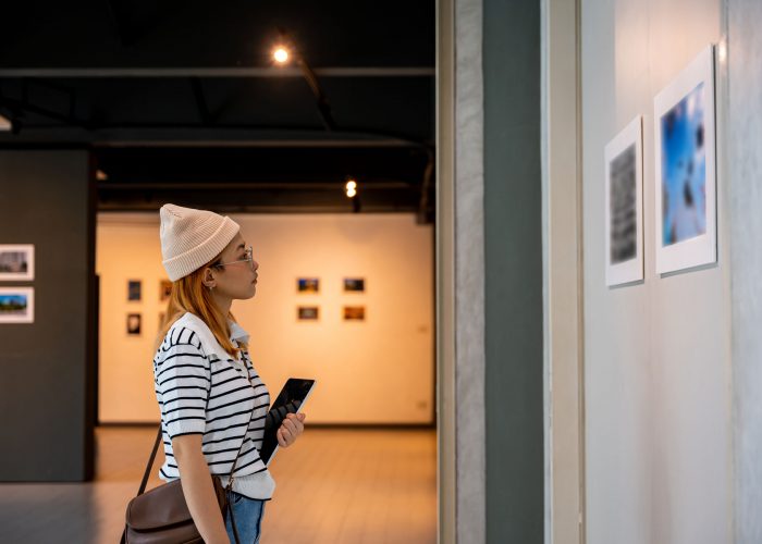 Young person at photo frame hold digital book leaning against at show exhibit artwork gallery, Asian woman holding tablet at art gallery collection in front framed paintings looking pictures on wall